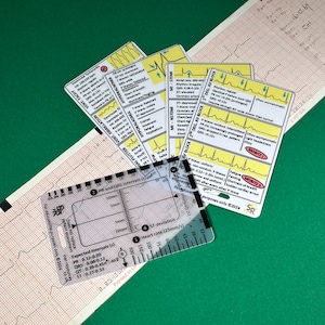 May include: A clear plastic ruler with black markings for measuring heart rate, ST deviation, and PR and QRS intervals. The ruler is labeled "for educational purposes only 2024".  The ruler is lying on top of a white sheet of paper with a grid pattern and a yellow and black card with a heart rate rhythm graphic and text that reads "2 DEG AV BLOCK".