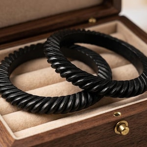 May include: Two black circular bangles with a twisted, rope-like texture resting inside a rectangular wooden jewellery box. The box features a hinged lid and a beige velvet interior with padded slots designed to hold jewellery securely in place.