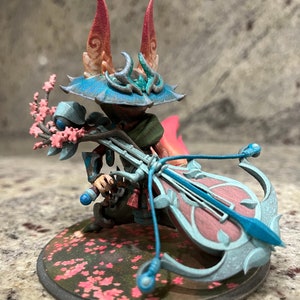 May include: A detailed figurine of a character with a blue hat and a musical instrument. The figure is holding a bow-shaped instrument with a pink and blue design. The base is decorated with pink flowers.