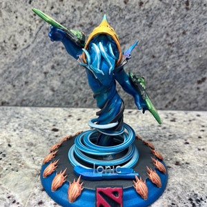 May include: A blue and green painted miniature figurine of a Dota 2 character,  a blue and white swirling base with a Dota 2 logo, and the word "Ionic" printed on the base.