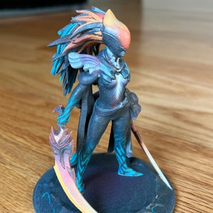 May include: A 3D printed figurine of a female warrior with a feathered headdress, holding two curved swords. The figurine is painted in shades of blue, orange, and black.
