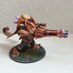 Puede incluir: Una figura en miniatura impresa en 3D de una criatura con un arma grande, que lleva una capa y un casco. La criatura está pintada en tonos de marrón, dorado y morado.