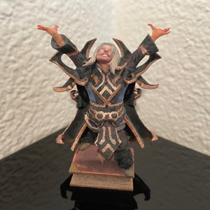 Op de afbeelding: Een 3D-geprint beeldje van een tovenaar met lang blond haar, gekleed in een zwarte en blauwe robe met gouden accenten. De tovenaar staat op een kleine, bruine, rechthoekige basis.