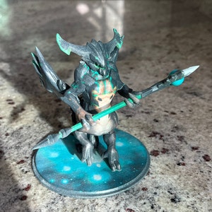 Puede incluir: Una miniatura impresa en 3D de una criatura negra y verde que sostiene un bastón con un orbe azul en el extremo. La criatura está de pie sobre una base redonda con un diseño de galaxia azul y blanco.