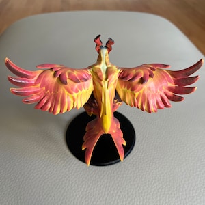 Puede incluir: Una figura de fénix roja y amarilla con las alas extendidas, de pie sobre una base negra. El fénix es una criatura mítica que simboliza el renacimiento y la renovación.