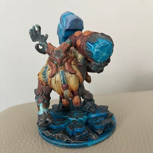 Peut inclure: Une figurine miniature peinte d'un nain tenant un grand marteau bleu recouvert de glace. Le nain porte une armure brune et dorée et a une longue barbe brune. La figurine est debout sur une base bleue et grise.