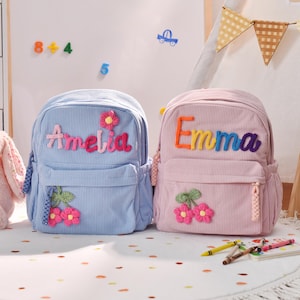 Peut inclure: Deux sacs à dos en velours côtelé, un bleu clair et un rose, chacun personnalisé avec un nom en lettres brodées colorées. Le sac bleu dit "Amelia" et le rose dit "Emma". Les deux ont des appliqués de fleurs et de feuilles.
