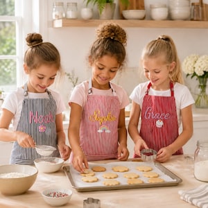 Peut inclure: Trois enfants portant des tabliers personnalisés en préparant des biscuits. Le tablier de gauche est bleu marine à rayures blanches avec le nom Noah, celui du centre est rouge à rayures blanches avec le nom Sophie, et celui de droite est rouge à rayures blanches avec le nom Grace.