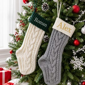 May include: Two knit Christmas stockings with cable patterns. One is cream-colored with a dark green cuff embroidered with the name Roman. The other is gray with a cream-colored cuff embroidered with the name Alex in gold-colored thread.