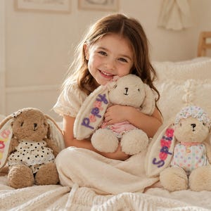 May include: Three plush rabbit toys with long floppy ears, each featuring embroidered names on the ear. One brown rabbit wears a white polka dot dress, one cream rabbit wears a pink dress, and one cream rabbit wears a multi-colored polka dot outfit.