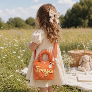 Könnte beinhalten: Eine kleine orangefarbene Cord-Handtasche mit dem Namen Freya in gelber Schreibschrift und einer kleinen weißen Hasen-Applikation. Die Tasche verfügt über zwei kurze Griffe, einen abnehmbaren orangefarbenen Schulterriemen und einen weißen Druckknopf.
