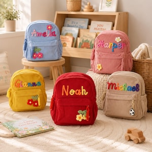 May include: Five corduroy backpacks in light blue, pink, yellow, red, and beige. Each bag features a name embroidered in colorful yarn, a front zippered pocket, and a small crocheted flower or soccer ball charm attached to the zipper pull.
