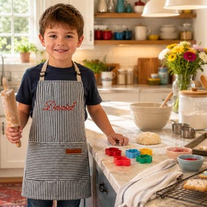 May include: A navy blue and white striped apron with a red embroidered script on the chest and a brown leather-like patch on the front pocket. The apron features adjustable neck straps and is worn over a dark blue t-shirt in a kitchen setting.