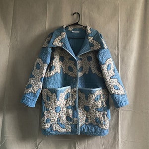 May include: A light blue quilted jacket with floral and leaf-shaped patches. The jacket has two front pockets and a button-up closure. The floral patches are white with small red, blue, and green flowers. The jacket is hanging on a black hanger.