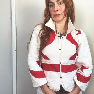 May include: A white jacket with red star and stripe designs. The jacket has a wide collar, brass-colored buttons, and three-quarter sleeves. The jacket is worn over a brown top and white pants.