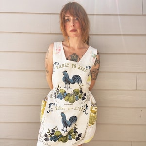 May include: A white apron with a rooster and fruit design. The apron has two pockets and says "Early to Rise, Birds to the Mist".