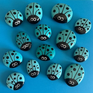 May include: A collection of hand-painted ladybug rocks in various sizes. Each rock is teal with black details, including spots and a ladybug face. The rocks are arranged on a bright blue surface.