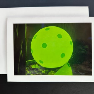 May include: A bright green, glow-in-the-dark, plastic pickleball with black dots, sitting on a bed of small, round, gray stones.