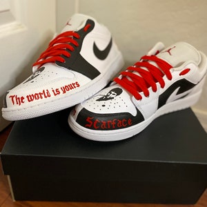 May include: White and black custom sneakers with red laces. The left shoe has the text "The world is yours" written on the side. The right shoe has the word "Scarface" written on the side.