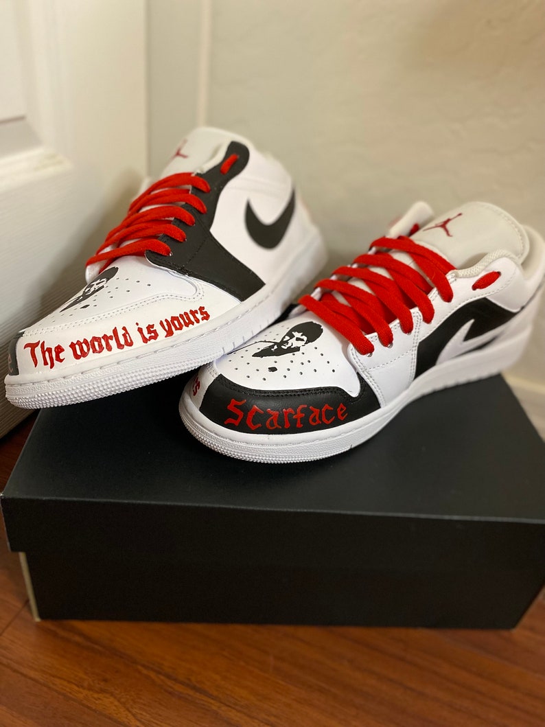 May include: White and black custom sneakers with red laces. The left shoe has the text "The world is yours" written on the side. The right shoe has the word "Scarface" written on the side.