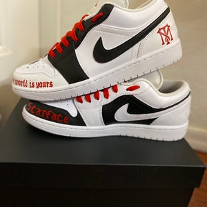 May include: A pair of white and black custom sneakers with red laces. The sneakers have the words "Scarface" and "The world is yours" written on them. The sneakers are sitting on top of a black shoebox.