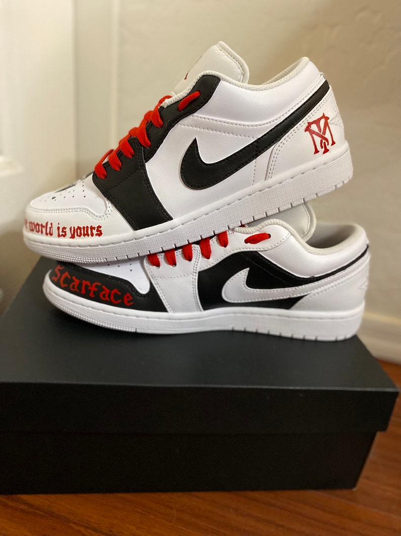 May include: A pair of white and black custom sneakers with red laces. The sneakers have the words "Scarface" and "The world is yours" written on them. The sneakers are sitting on top of a black shoebox.