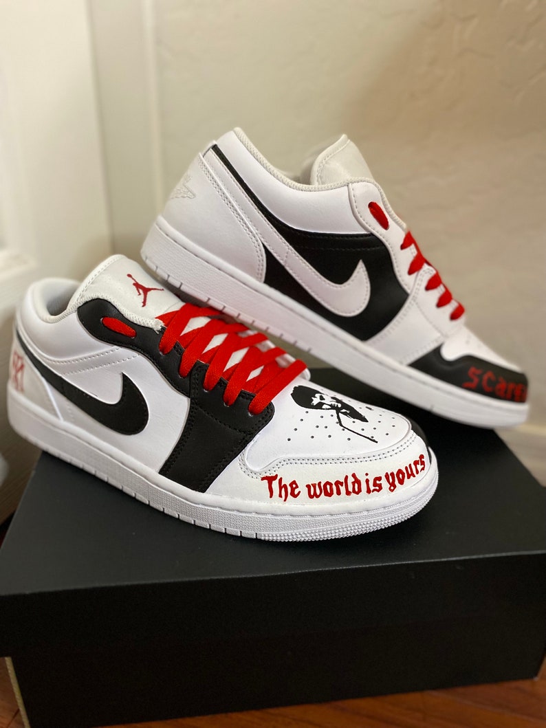 May include: White and black low-top sneakers with red laces. The right shoe has the text "The world is yours" written on the side. The left shoe has the text "5 Cars" written on the side.