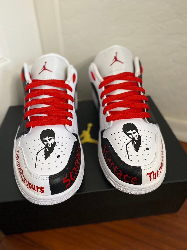 May include: A pair of white and black sneakers with red laces. The sneakers have a black and white design with the words "Scarface" and "The world is yours" written on them. The sneakers also feature a portrait of Al Pacino as Tony Montana.