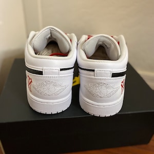 May include: A pair of white leather low-top sneakers with black accents and red stitching. The sneakers have a white midsole and a black outsole. The Jordan logo is visible on the tongue and the back of the shoe.
