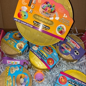 Puede incluir: Una colección de sets de juguetes misteriosos Squishy Bun, cada uno con un recipiente de vaporera de plástico amarillo redondo con una etiqueta de cartón colorida. Las etiquetas muestran ilustraciones de pequeños personajes de albóndigas arcoíris y texto que incluye Squishy Bun, Sugar Edition y 3+.