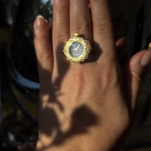 May include: A gold-colored ring with a small, round watch face. The ring has an ornate, filigree design around the watch. The watch face is light blue with white hands and markers. The ring is on a person's hand.