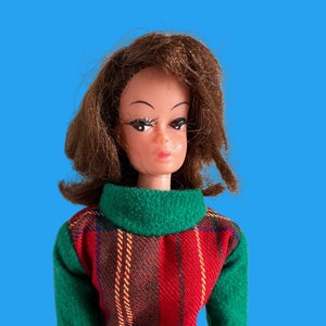May include: A vintage doll with brown hair and a red plaid dress with green sleeves and a green collar. The doll has a fair complexion and is set against a blue background.