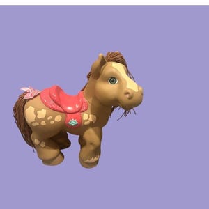 May include: A tan toy horse with a red saddle and a pink bow on its tail. The horse has brown hair and spots on its body. The horse has green eyes and a white patch on its face. The toy is set against a light purple background.