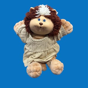 Può includere: Una bambola di peluche vintage con un viso rotondo, occhi blu e un naso marrone. La bambola ha capelli di filo marrone con un ciuffo bianco e indossa un abito a maglia beige. Le braccia e le gambe della bambola sono color cuoio.