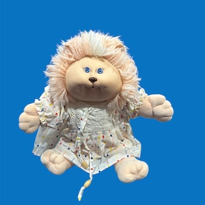 Peut inclure: Une poupée en peluche vintage avec une crinière de lion et un visage rond. La poupée a des yeux bleus et porte une robe blanche à motifs colorés. La crinière de la poupée est rose clair.