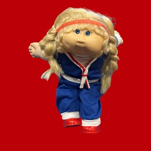Puede incluir: Muñeca vintage con cabello rubio, ojos azules y diadema roja. La muñeca lleva un atuendo azul con ribetes blancos y rojos, y zapatos rojos. Un juguete coleccionable clásico.