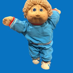 Peut inclure: Une poupée Cabbage Patch Kid vintage, avec un corps doux et un visage rond avec des yeux bleus. La poupée porte un survêtement bleu avec des détails rouges et blancs, et des baskets blanches. La poupée a des cheveux bouclés brun clair.