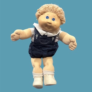 Peut inclure: Une poupée vintage avec un visage rond, des yeux bleus et des cheveux blonds bouclés. La poupée porte une chemise blanche à col, une salopette en jean, des chaussettes blanches et des chaussures blanches. Les bras de la poupée sont tendus, sur fond bleu sarcelle.