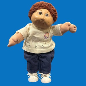 May include: A vintage Cabbage Patch Kid doll with brown yarn hair, a cream-colored sweater, dark blue jeans, and white sneakers with blue stripes. The doll has a round face and is posed with arms outstretched.