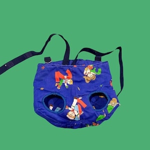 May include: A blue baby walker with cartoon figures in red, green, and yellow. The walker has two leg holes and adjustable straps. The background is a solid green.