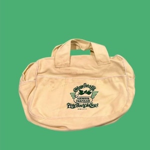 Peut inclure: Sac fourre-tout en toile crème avec deux poignées et un logo vert. Le logo comporte le texte "Cabbage Patch Kids" au-dessus de "Airways Transair" et "P'tits Bouts de Chou". Le sac est de forme arrondie et possède une poche latérale.