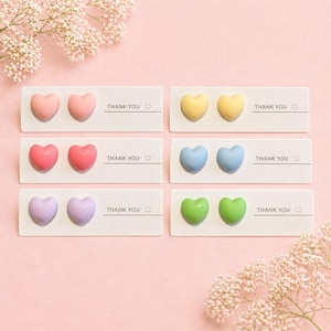 May include: Six cards, each with two heart-shaped decorations in pastel shades of pink, yellow, red, blue, purple, and green. The cards read "THANK YOU" with a smiling face. A floral arrangement is in the background.