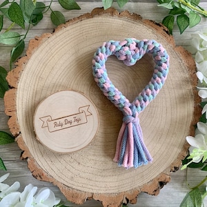May include: A heart-shaped dog toy made of braided rope in pink, blue and green colours. The toy has a tassel at the bottom and is displayed on a wooden slice with the text "Ruby Dog Toys" engraved on it.