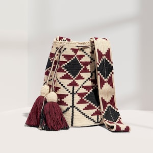 May include: A crocheted bag with a geometric pattern in shades of beige, burgundy and black. The bag has a long strap and two burgundy tassels.
