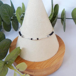 Peut inclure: Un bracelet argenté délicat avec des perles noires et argentées. Le bracelet est présenté sur un présentoir conique blanc avec une base en bois, entouré de verdure. Le bracelet comporte une chaîne argentée texturée.