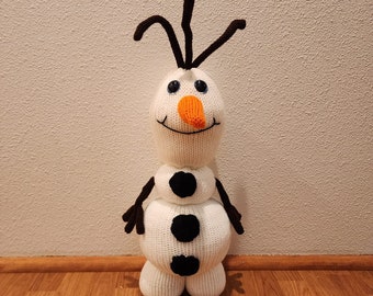 May include: A white knitted snowman with black eyes, a black mouth, and three black buttons. The snowman has an orange carrot nose and black yarn hair.