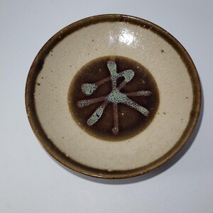 Pode incluir: Uma tigela de cerâmica redonda com interior creme salpicado e borda marrom escura. O centro apresenta uma mancha circular marrom escura com um design azul claro em forma de estrela ou cruz abstrata pintado com esmalte texturizado.