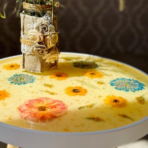 May include: A white round side table with wooden legs features a yellow resin tabletop with embedded dried flowers and gold glitter.