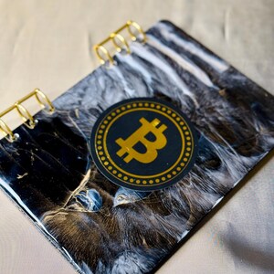 May include: A rectangular notebook with a black, white, and gold marbled resin cover. The notebook features gold-colored metal spiral binding and a circular black sticker with a gold Bitcoin symbol centered on the front cover.