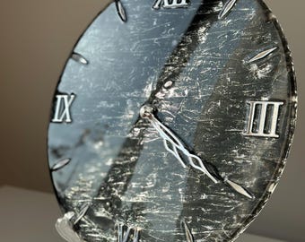 May include: A black and silver clock with Roman numeral markings on a clear glass face. The clock is on a black stand with a clear plastic base.